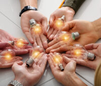 Mehrere Menschen halten kreisförmig ihre Hände nebeneinander auf einer Unterlage und es liegen leuchtende Glühbirnen in den Händen als Zeichen für Energy Sharing.