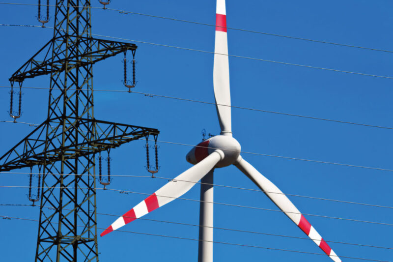 Windenergieanlage der Marke Enercon hinter einer Stromleitung mit langer Brennweite (Teleobjektiv) fotografiert. Symbolbild für CfD