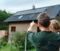 Ein Mann mit Kind im Arm vor Haus mit Photovoltaik-Anlage.