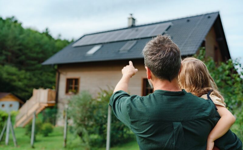 Ein Mann mit Kind im Arm vor Haus mit Photovoltaik-Anlage.