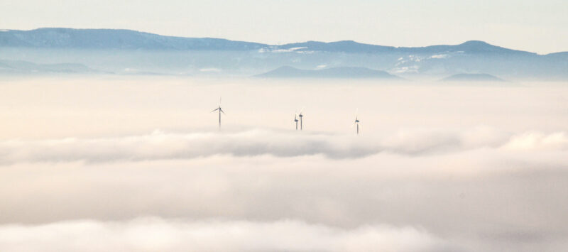 Eine Wolkendecke, aus der die Spitzen von Windenergieanlagen etwas verschwommen herausragen. Im Hintergrund bewaldete Berghänge.