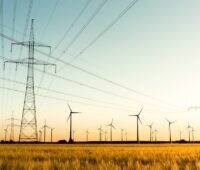 Strommasten auf einem Feld, im Hintergrund Windkraftanlagen - Versorgungssicherheit auch mit erneuerbaren Energien.