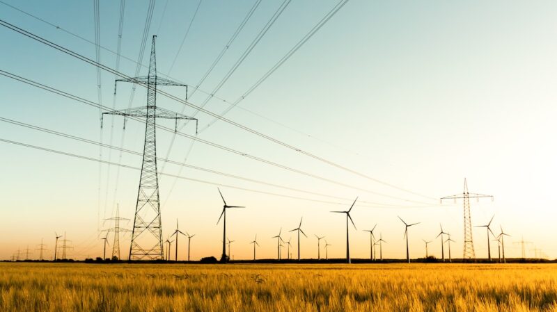 Strommasten auf einem Feld, im Hintergrund Windkraftanlagen - Versorgungssicherheit auch mit erneuerbaren Energien.