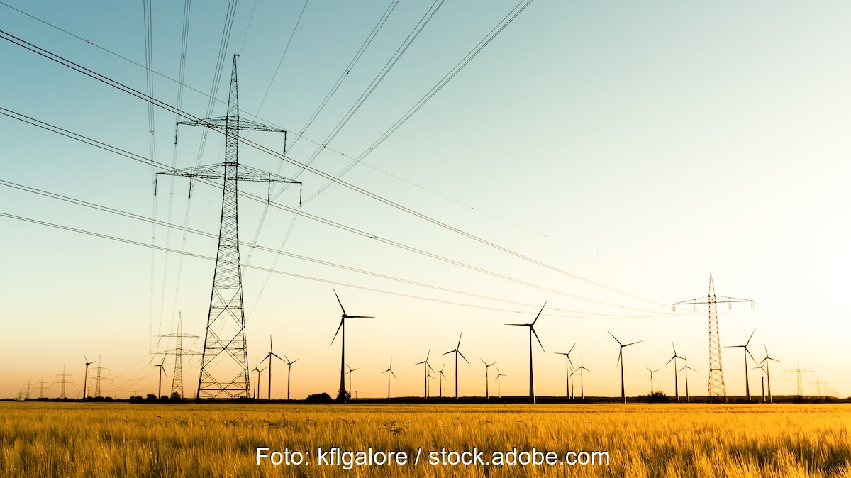 Strommasten auf einem Feld, im Hintergrund Windkraftanlagen - Versorgungssicherheit auch mit erneuerbaren Energien.