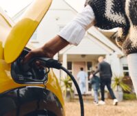 Im Vordergrund verbindet eine Person ein Ladekabel mit einem Elektroauto. Im Hintergrund gehen ein Mann und zwei Kinder in ein Einfamilienhaus. Symbol für das Thema: Sparen mit dynamischen Stromtarifen.