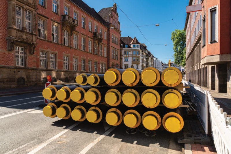 Ein Stapel Fernwärmeleitung liegt zwischen Gebäuden auf einer Straße.