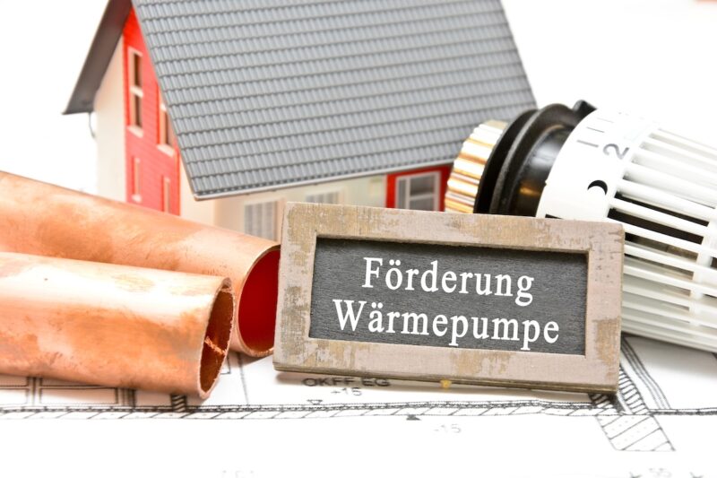 Symbolbild: im Vordergrund eine Tafel mit der Aufschrift "Förderung Wärmepumpe" - in Deutschland läuft die über die BEG -, rechts Thermostat, links Heizungsrohre, im Hintergrund das Modell eines Hauses