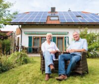 Ein Paar sitzt im Garten vor einem Einfamilienhaus mit Photovoltaikanlage auf dem Dach.