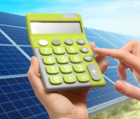 Hände mit Taschenrechner vor einer Freiflächen-Solaranlage.