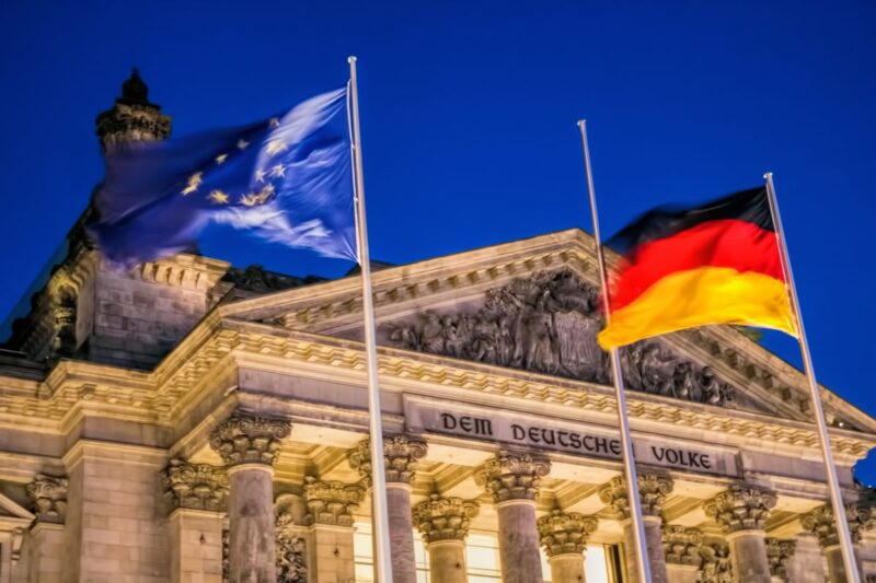 Beleuchtetes Gebäude des Deutschen Bundestages mit Europa- und Deutschlandflagge im Vordergrund.