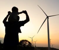 Vater mit Kind im Gegenlicht vor Windenergieanlagen bei Sonnenaufgang - Symbol für Akzeptanz von Windkraft