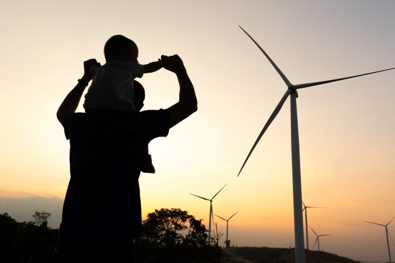 Kind auf den Schultern eines Mannes vor Windkraftanlage bei Sonnenuntergang.