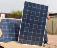 Person hält ein großes PV-Modul neben einem Karton mit weiteren PV-Komponenten.