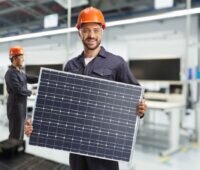 Arbeiter mit Helm präsentiert PV-Modul in Fabrikhalle.