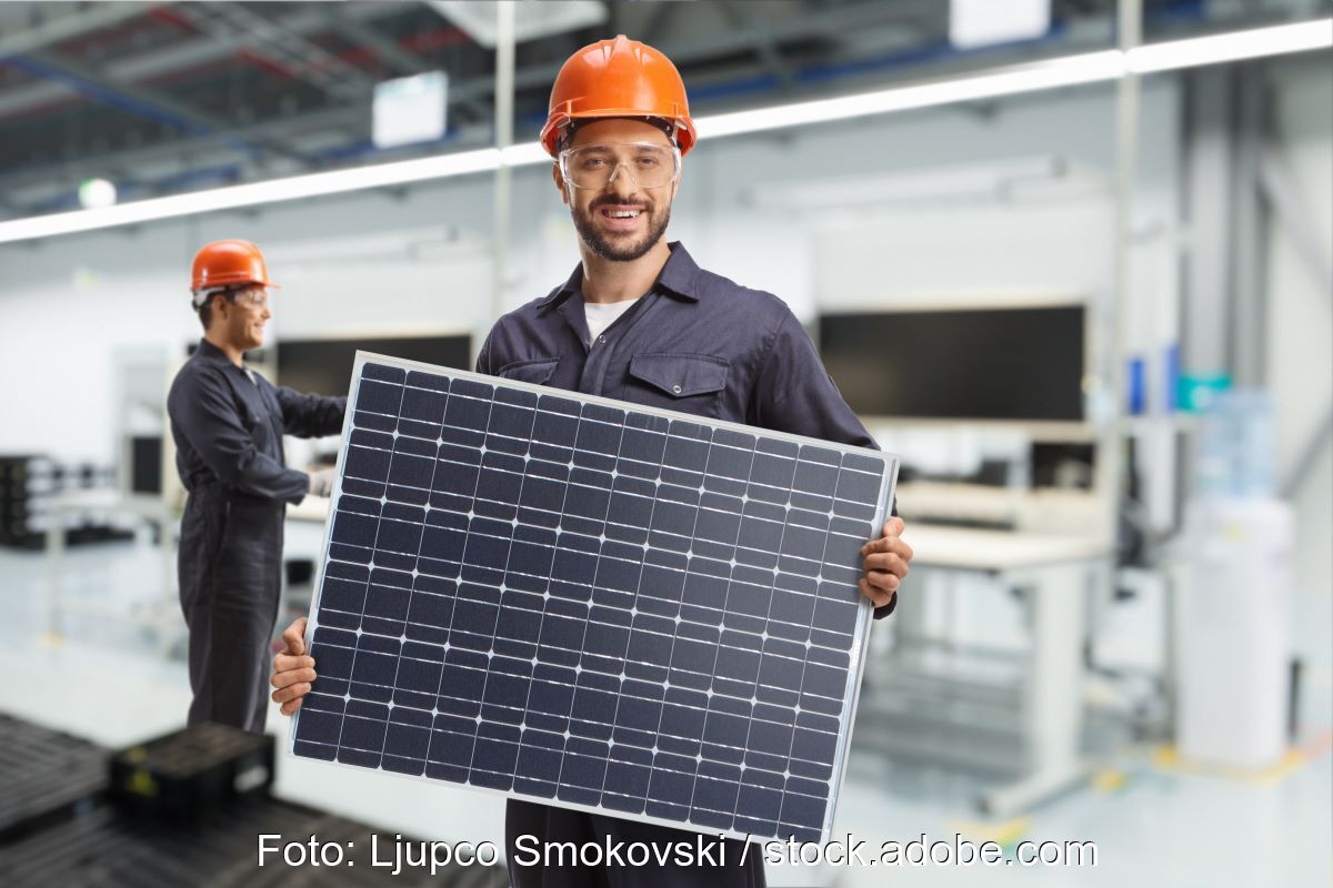 Arbeiter mit Helm präsentiert PV-Modul in Fabrikhalle.