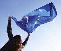 Junge Frau schwenkt Europafahne vor blauem Himmel. Symbol für Made in Europe.
