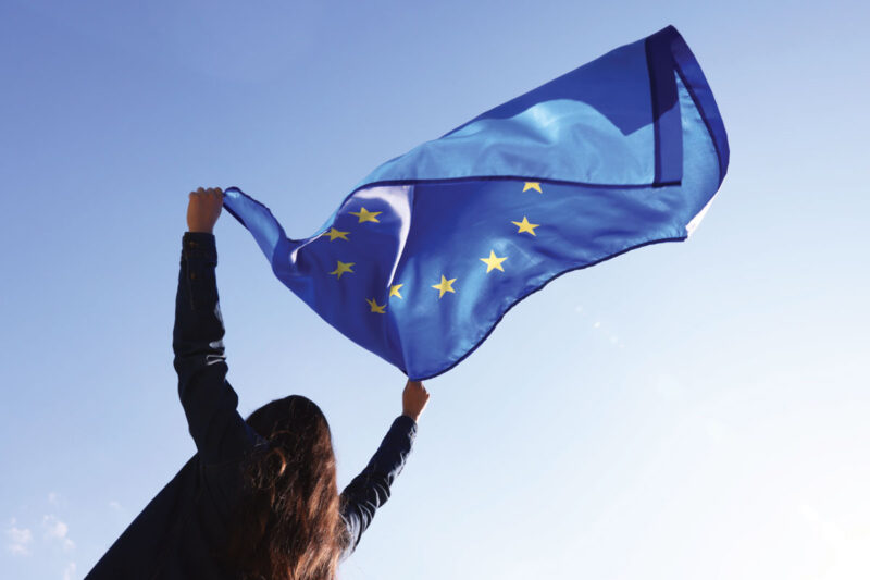 Junge Frau schwenkt Europafahne vor blauem Himmel. Symbol für Made in Europe.