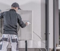 Ein Monteur installiert oder deinstalliert eine Wärmepumpe an bzw. von einer Häuserwand.
