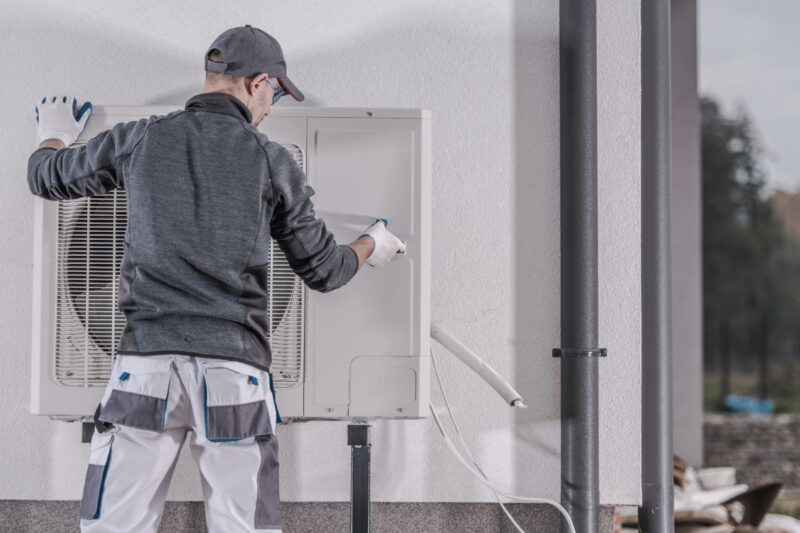 Ein Monteur installiert oder deinstalliert eine Wärmepumpe an bzw. von einer Häuserwand.