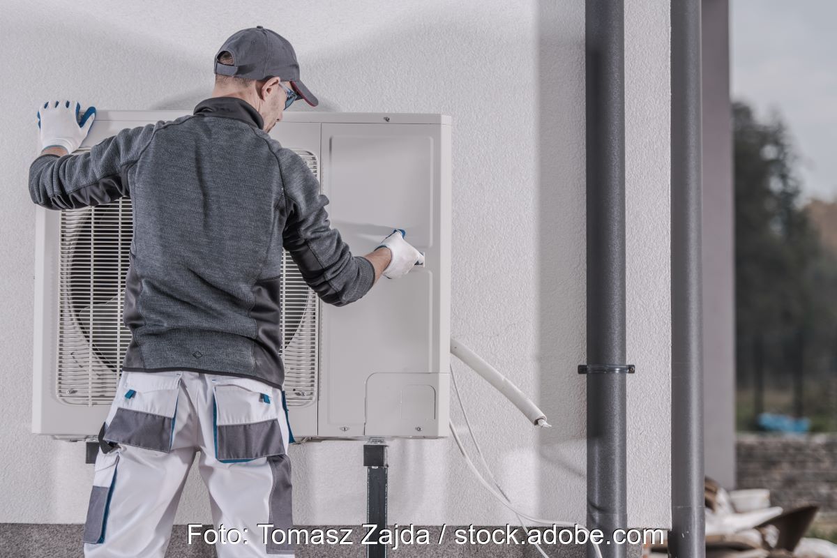 Ein Monteur installiert oder deinstalliert eine Wärmepumpe an bzw. von einer Häuserwand.