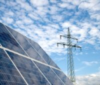 Freiflächen-Solarmodul udn Hochspannungsmast unter blauem Himmel mit Wolken.