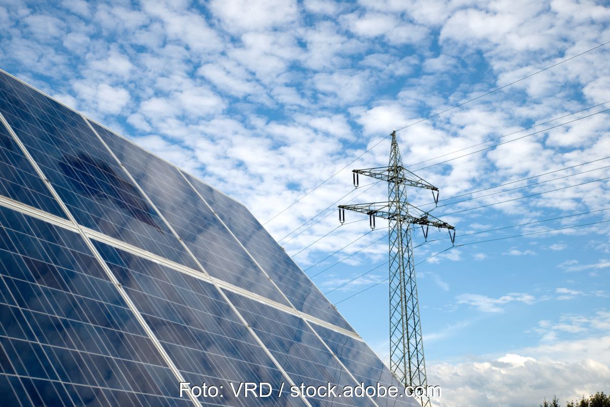 Freiflächen-Solarmodul udn Hochspannungsmast unter blauem Himmel mit Wolken.