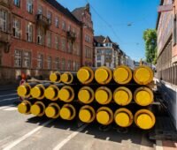 Stapel von Rohren zum Wärmetransport auf einer Straße in einer Stadt mit historischen Gebäuden.