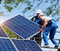 Zwei Monteure platzieren bei Sonnenschein ein Solarmodul auf ein Schrägdachgestell.