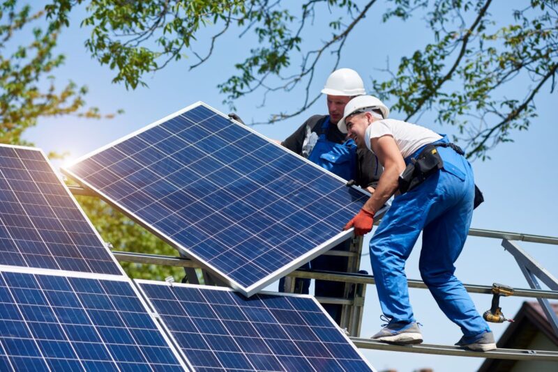 Zwei Monteure platzieren bei Sonnenschein ein Solarmodul auf ein Schrägdachgestell.