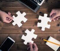 Fokus auf drei große Puzzleteile, von Fingern gehalten und Taschenrechnern und Computern.