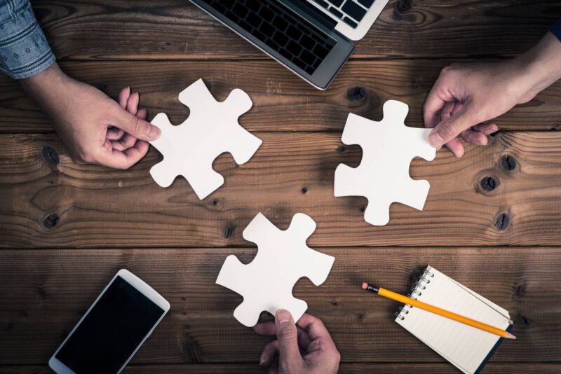 Fokus auf drei große Puzzleteile, von Fingern gehalten und Taschenrechnern und Computern.