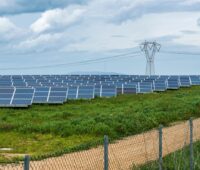 Ein eingezäuntes Feld mit PV und einem Hochspannungsmast.