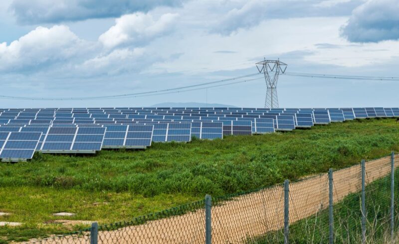 Ein eingezäuntes Feld mit PV und einem Hochspannungsmast.