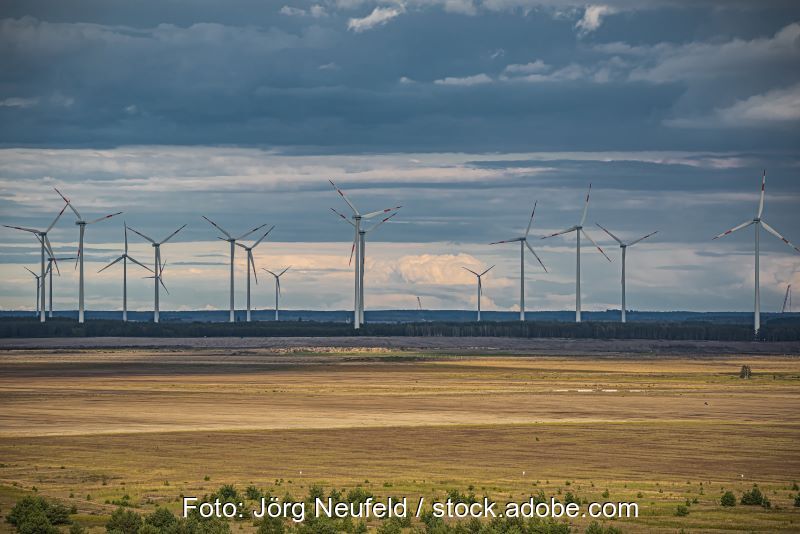 Windräder in flacher Landschaft auf braunem Boden vor Wolken.