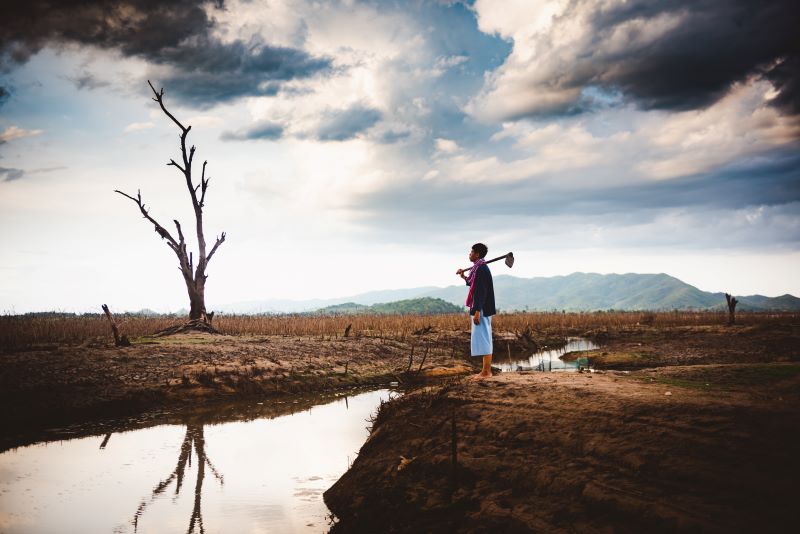 Ein asiatischer landwirt blickt auf vertrockneten Baum und Land mit wenig Wasser.