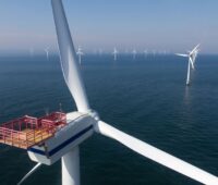 Blick über einen Offshore-Windpark aus der Vogelperspektive, im Vordergrund links eine Windturbine und Rotoren.