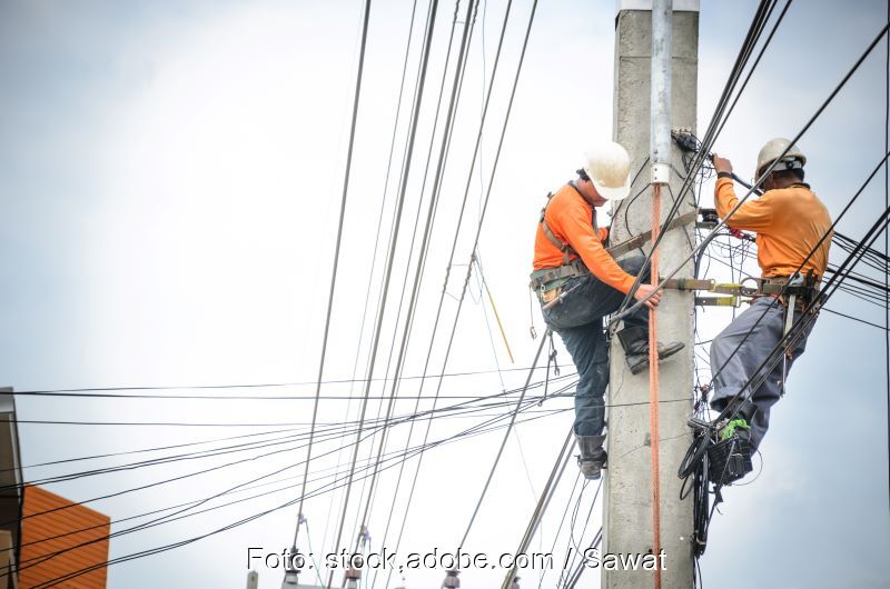 Zwei Arbeiter auf einem Strommast mit Leitungen in vielen Richtungen.