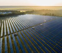 Luftaufnahme von schräg vorn von einem großen Freiflächen-Solarpark bei tiefstehender Sonne, eingegrenzt von Bäumen und grünen Flächen im Hintergrund.