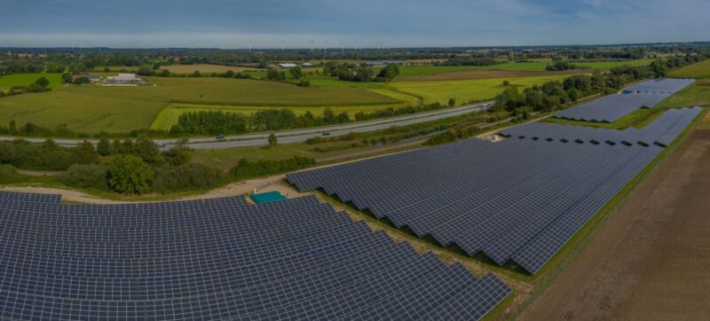 Freiflächen-Solarpark im Flachland an der Autobahn, am Horizont Windanlagen.