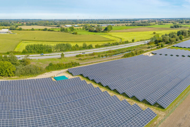 Freiflächen-Solarpark im Flachland an der Autobahn, am Horizont Windanlagen.