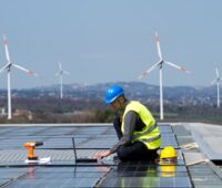 Ein Handwerker montiert Solarmodule auf einem Flachdach. Im Hintergrund drehen sich SWindräder.