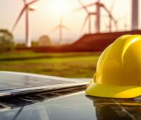 Gelber Arbeitshelm liegt auf Solarmodulen mit Windenergieanlagen im Hintergrund, zweiseitige Differenzverträge für künftige Photovoltaik und Windenergie sollten einen Marktwertkorridor berücksichtigen.