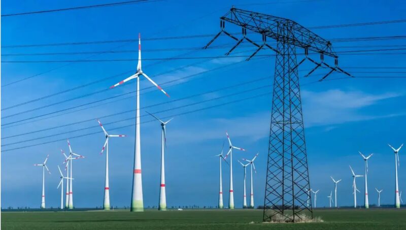 Mehrere Windenergieanlagen hinter Strommast, vor einem blauem Himmel mit leichten Schlieren.