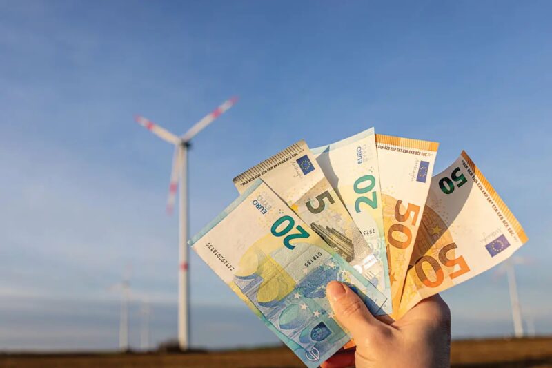 Eine Hand hält Geldscheine vor Windenergieanlage, die Redispatch-Abrechnung erfolgt ab 2026 direkt zwischen Anlagenbetreiber und Netzbetreiber.