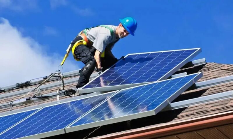 Das Foto zeigt einen Monteur in Arbeitskleidung, gesichert durch einen Gurt und mit blauem Schutzhelm auf dem Kopf, auf einem roten Dach vor blauem Himmel, der ein PV-Modul installiert.