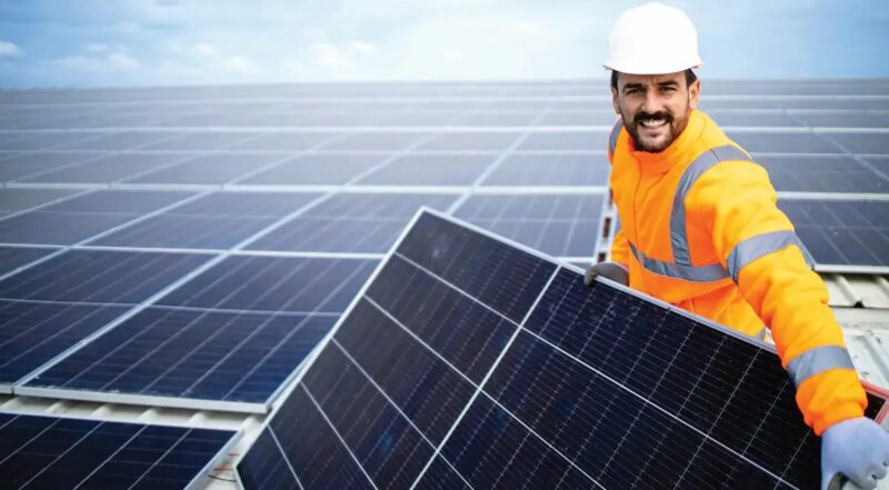 Ein Mann in oranger Arbeitskleidung mit weißem Schutzhelm hält ein Solarmodul vor einem Schrägdach mit weiteren Modulen in den Händen.
