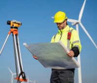 Ein Mann in Arbeitskleidung hält einen Plan vor Windenergieanlagen in den Händen, der TÜV hat den Windenergie-Ertragsgutachter Anemos-Jacob übernommen.