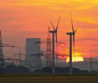 Windenergieanlagen, Strommasten und ein fossiles Kraftwerk liegen in einem orange-roten Sonnenuntergang.