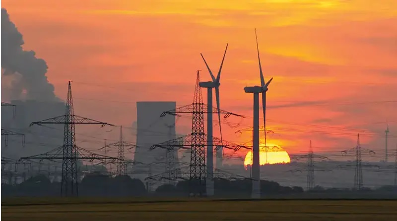 Windenergieanlagen, Strommasten und ein fossiles Kraftwerk liegen in einem orange-roten Sonnenuntergang.