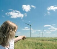 Eine junge Frau zeigt Windkraftanlagen auf einer grünen Wiese vor blauem Himmel als Zeichen für Bürgerenergie.